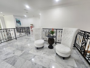 Vestíbulo con suelo de mármol, dos sillones blancos tapizados, una mesa decorativa con una planta y una barandilla de hierro forjado negro.