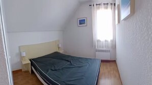Une petite chambre avec un lit simple, un plancher en bois, une tête de lit blanche et une fenêtre avec des rideaux.