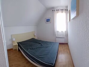 Ein kleines Zimmer mit einem Einzelbett, Holzboden, einem weißen Kopfteil und einem Fenster mit Vorhängen.