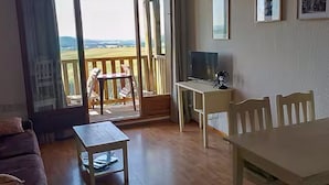 Une pièce dotée d’une porte-fenêtre coulissante donnant sur un balcon, d’une petite table, d’un téléviseur posé sur un meuble et de quelques chaises.