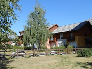 A traditional chalet-style building with a sloped roof, wooden balconies, and a fenced yard with rocks and greenery. - Semi-detached chalet south, shuttle to slope - 7 guests (La Salle-les-Alpes)