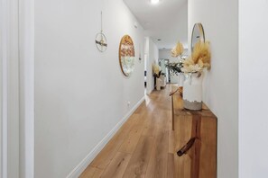 A hallway with wooden flooring, white walls, and decorative wall art including a circular mirror and a wall-mounted light fixture. - Boater’s Retreat | 4BR Home Near the Beach (Sebastian)
