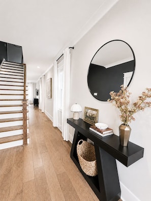 Un pasillo moderno con escaleras de madera, una consola negra con espejo y un jarrón con flores secas.