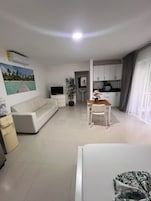 A hotel room with a bed, a sofa, a dining table with chairs, a TV, a potted plant, a kitchen area, and a bathroom.
