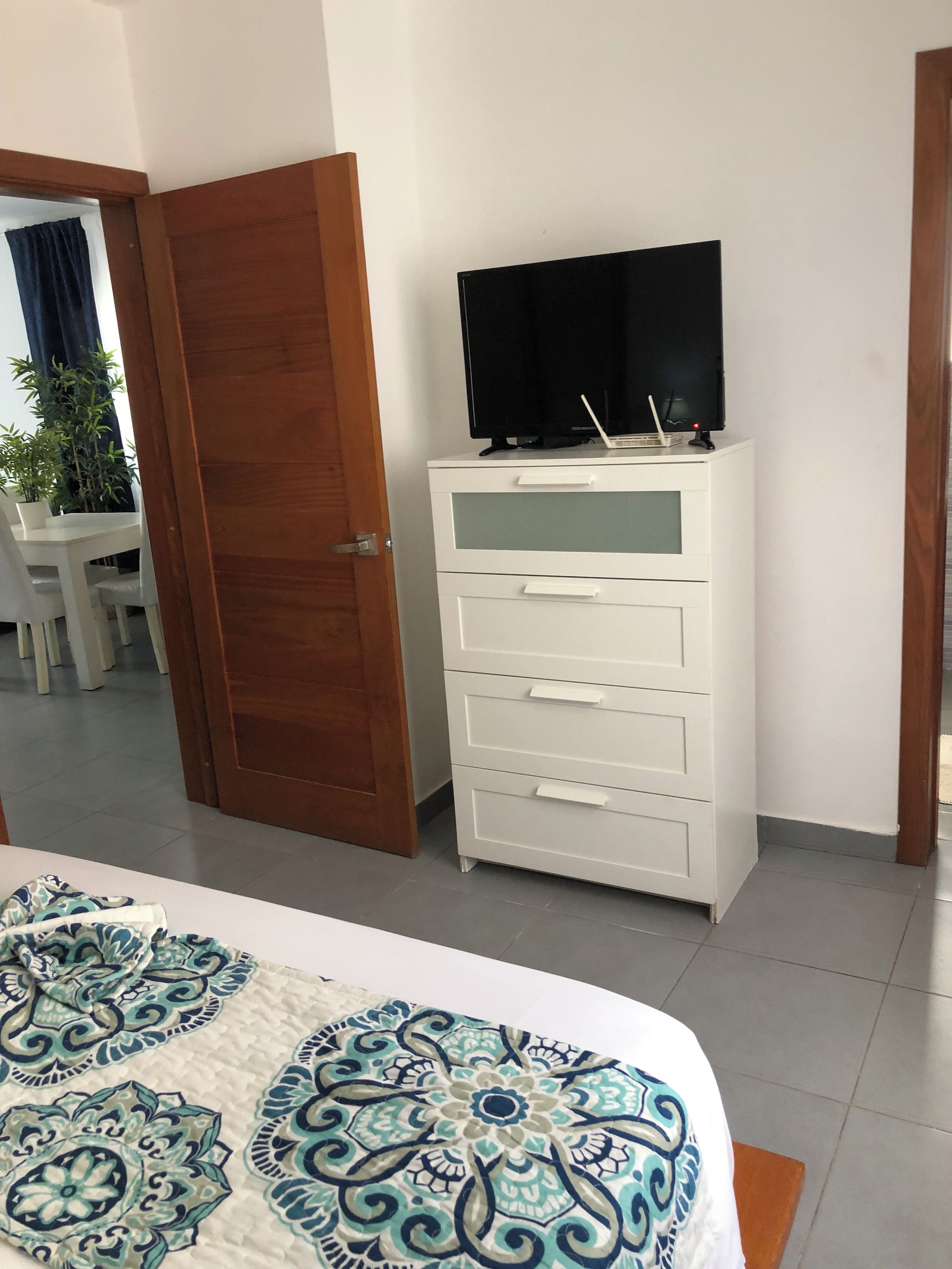 Ein Hotelzimmer mit einem Bett, einer weißen Kommode mit Schubladen, einem Fernseher darauf und einer Tür zu einem anderen Raum.