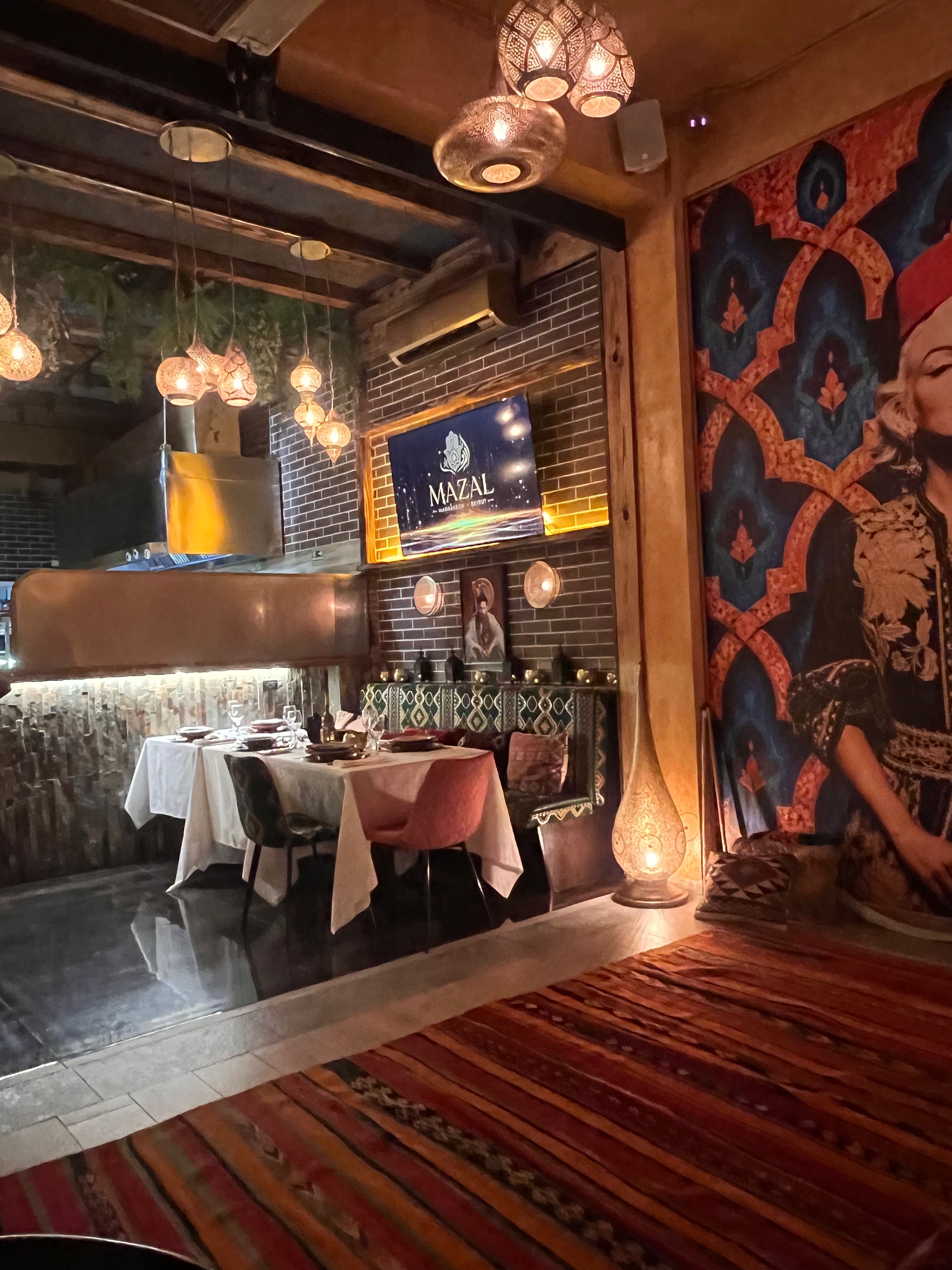 Et restaurantinteriør med hængende lys, en murstensvæg, et muralmaleri og et skilt med teksten 'MAZAL'.