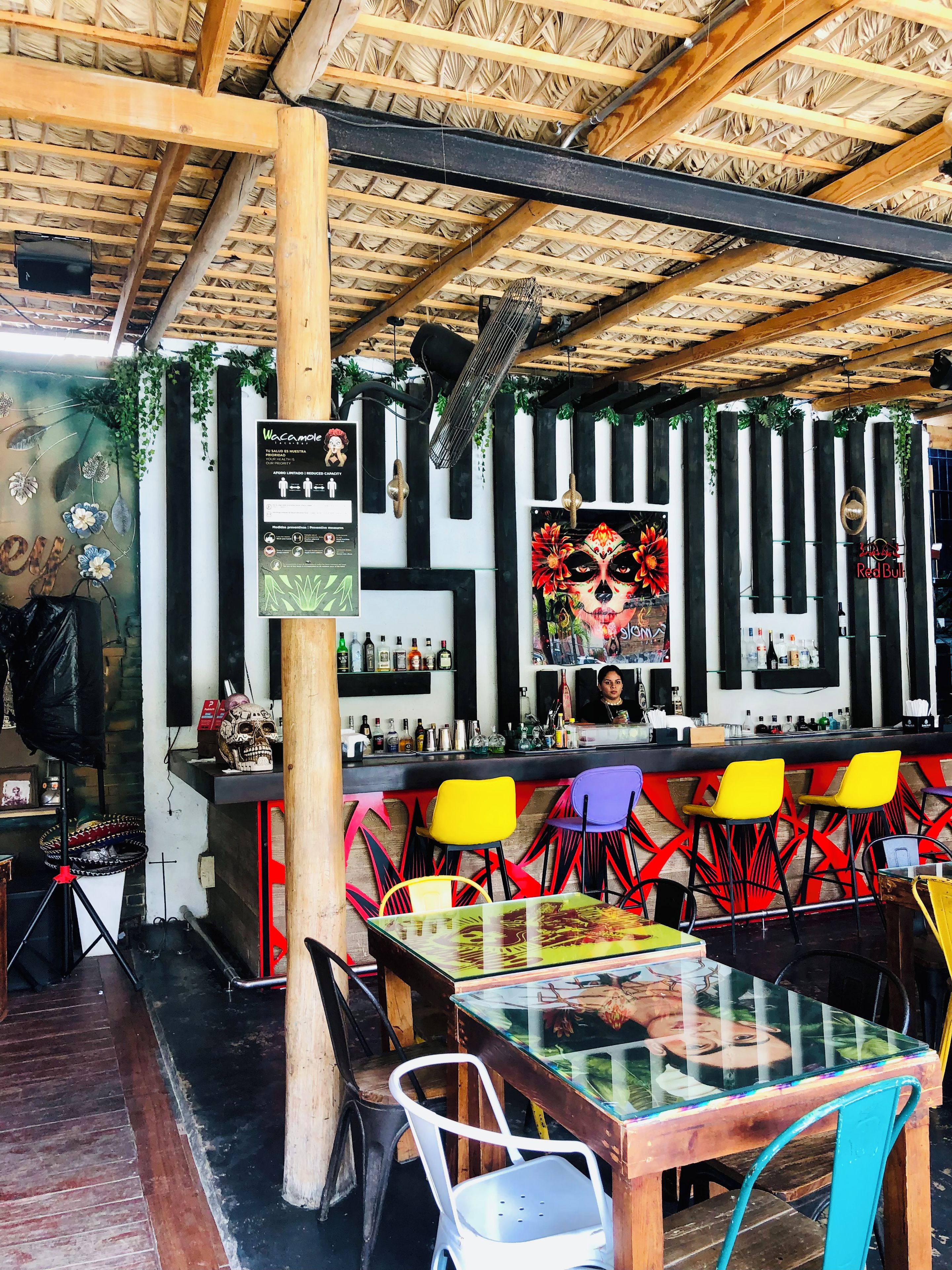 Um bar interno com teto de palha, vigas de madeira e diversos cadeiras e mesas coloridas.