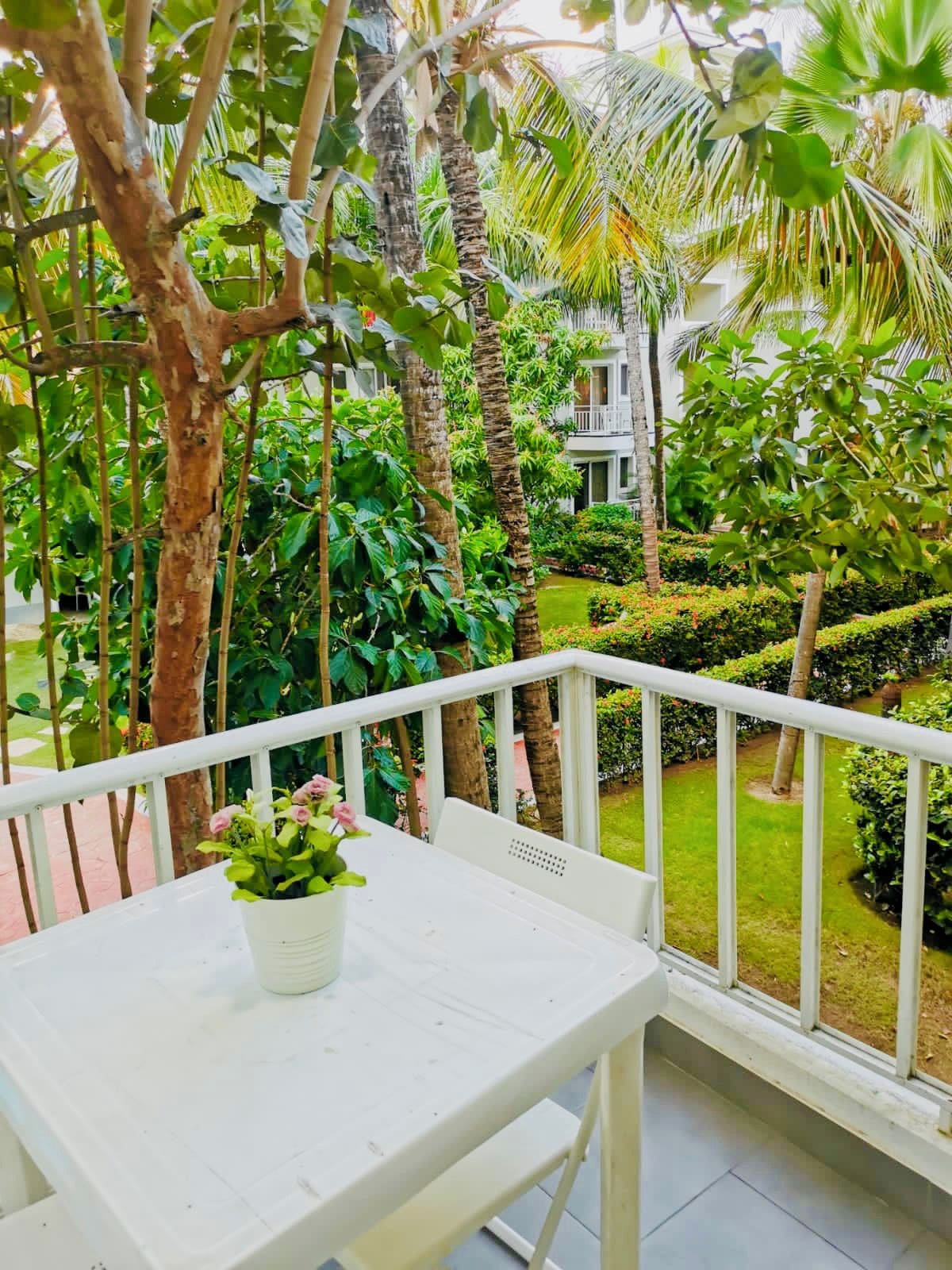 A white outdoor seating area with a table and a potted plant, surrounded by lush greenery and a well-maintained garden.