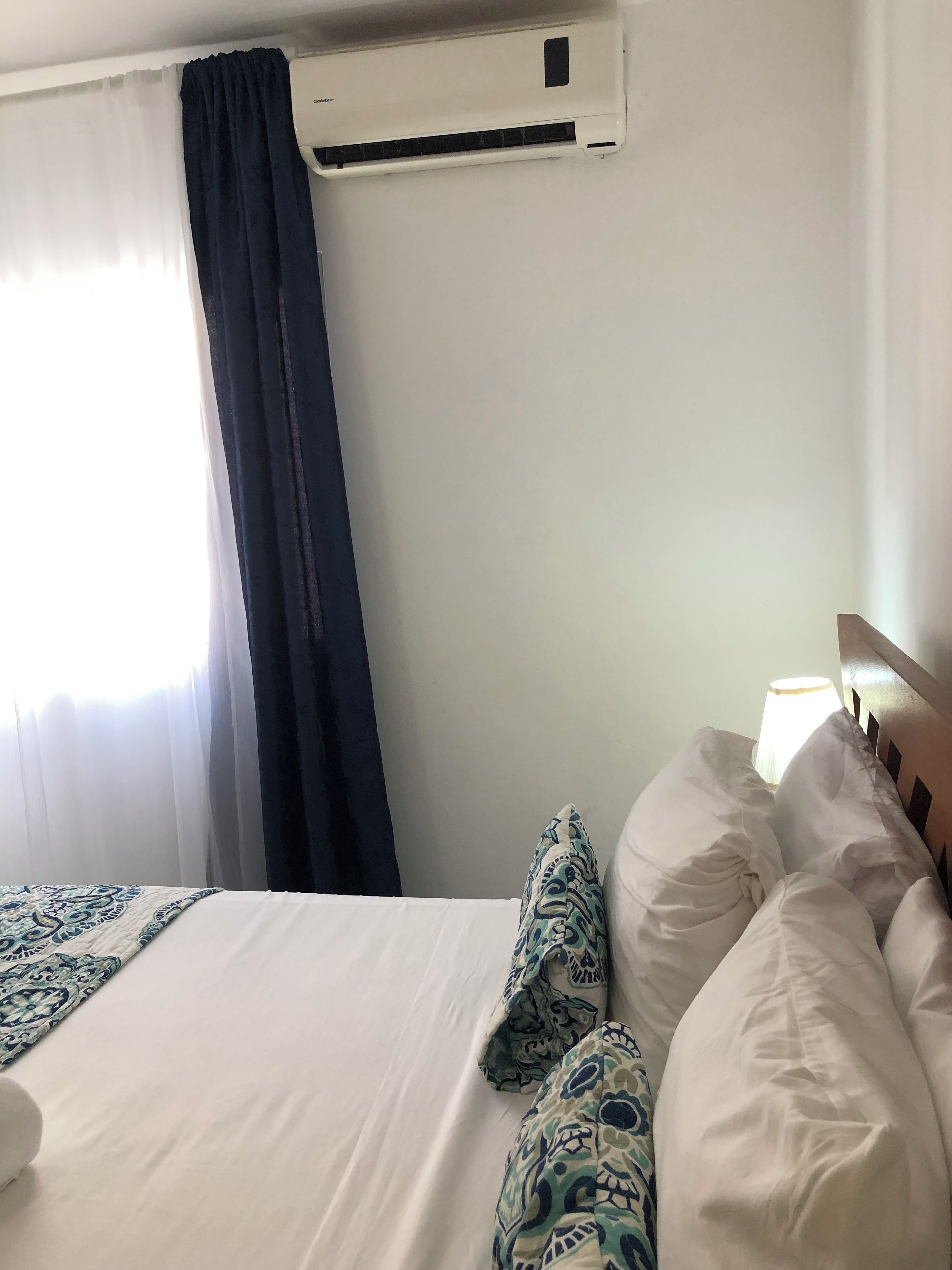 A hotel room with a bed, a window with curtains, an air conditioning unit, and a bedside lamp.