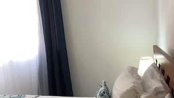A hotel room with a bed, a window with curtains, an air conditioning unit, and a bedside lamp.