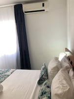 A hotel room with a bed, a window with curtains, an air conditioning unit, and a bedside lamp.