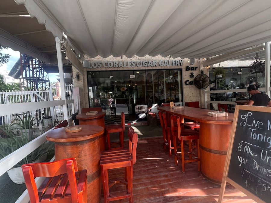 Un espace extérieur aménagé avec des tables et des chaises en bois, un comptoir de bar et une enseigne indiquant l’entrée de la galerie de cigares Los Corales.