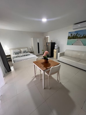 Una habitación de hotel con una cama, una mesa de comedor con sillas, un sofá blanco, un refrigerador y un cuadro en la pared.