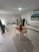 A hotel room with a bed, a dining table with chairs, a white sofa, a refrigerator, and a painting on the wall.