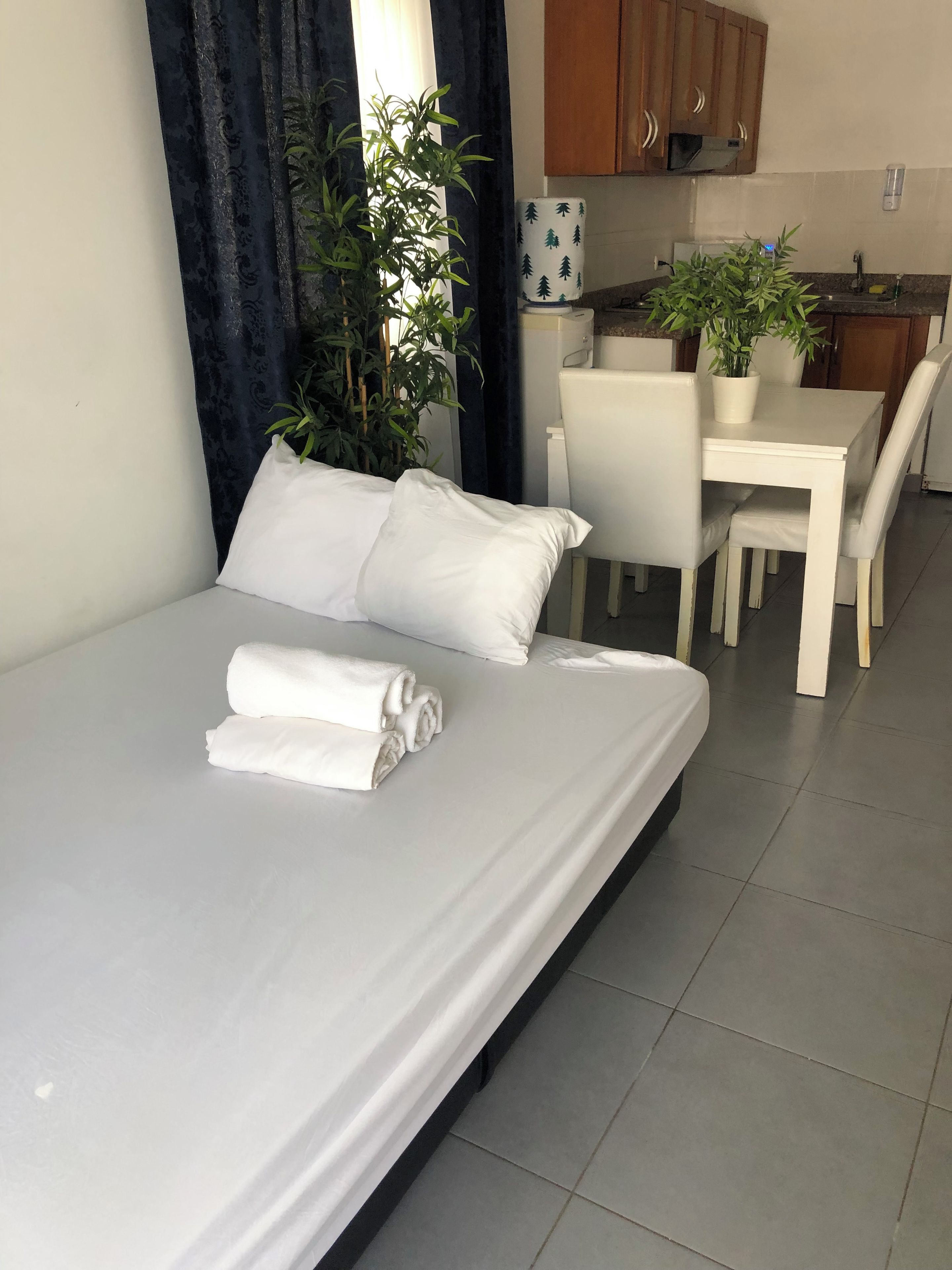 Una cama bien hecha con ropa de cama blanca, una mesa de comedor con sillas y una planta en maceta al fondo.