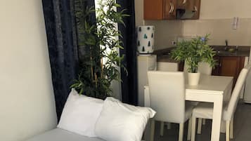 Una cama bien hecha con ropa de cama blanca, una mesa de comedor con sillas y una planta en maceta al fondo.