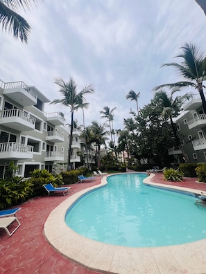 A swimming pool with lounge chairs, surrounded by palm trees and a multi-story building with balconies. - Sol Caribe Tropical Beach Suites (Punta Cana)