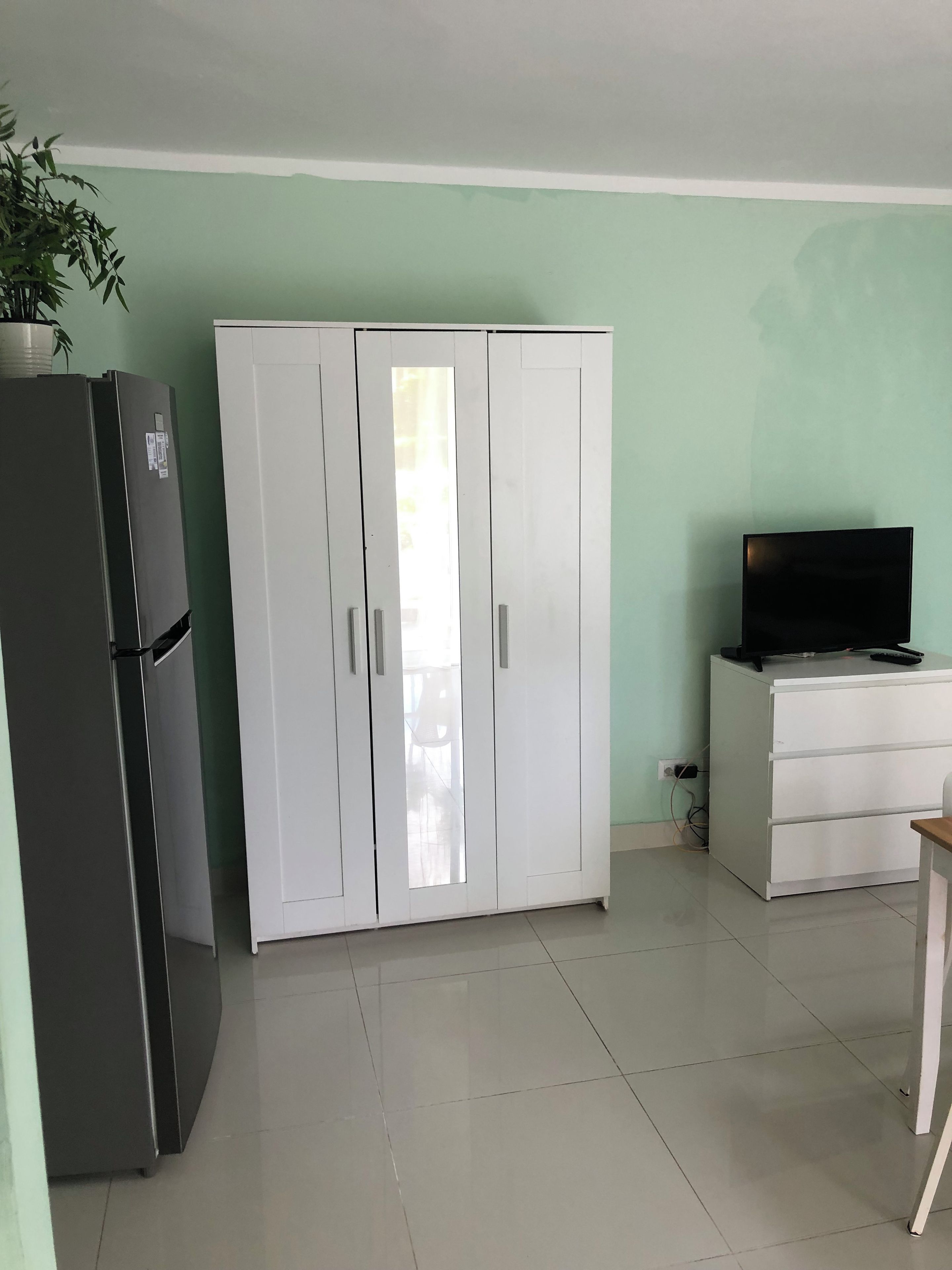 Una habitación con un armario blanco, un refrigerador negro, un escritorio blanco con televisor y una planta en maceta.