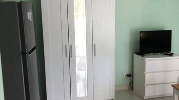 Una habitación con un armario blanco, un refrigerador negro, un escritorio blanco con televisor y una planta en maceta.