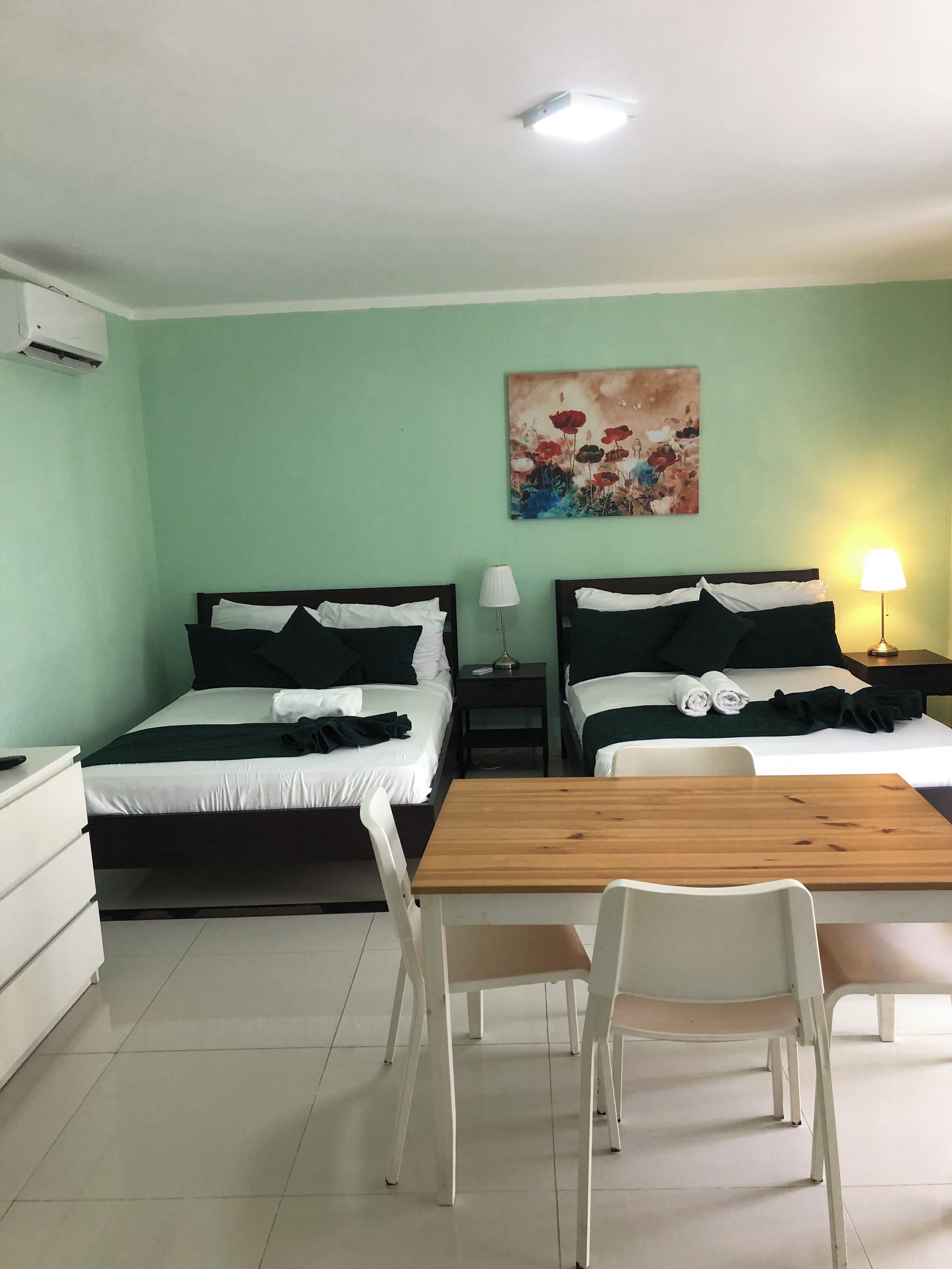 Quarto de hotel com duas camas, uma mesa de madeira, cadeiras brancas, um quadro na parede e uma luminária na mesinha de cabeceira.