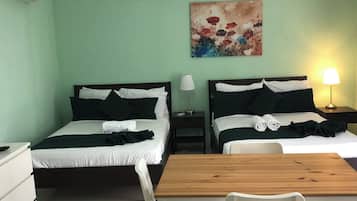Habitación de hotel con dos camas, una mesa de madera, sillas blancas, un cuadro en la pared y una lámpara en la mesita de noche.