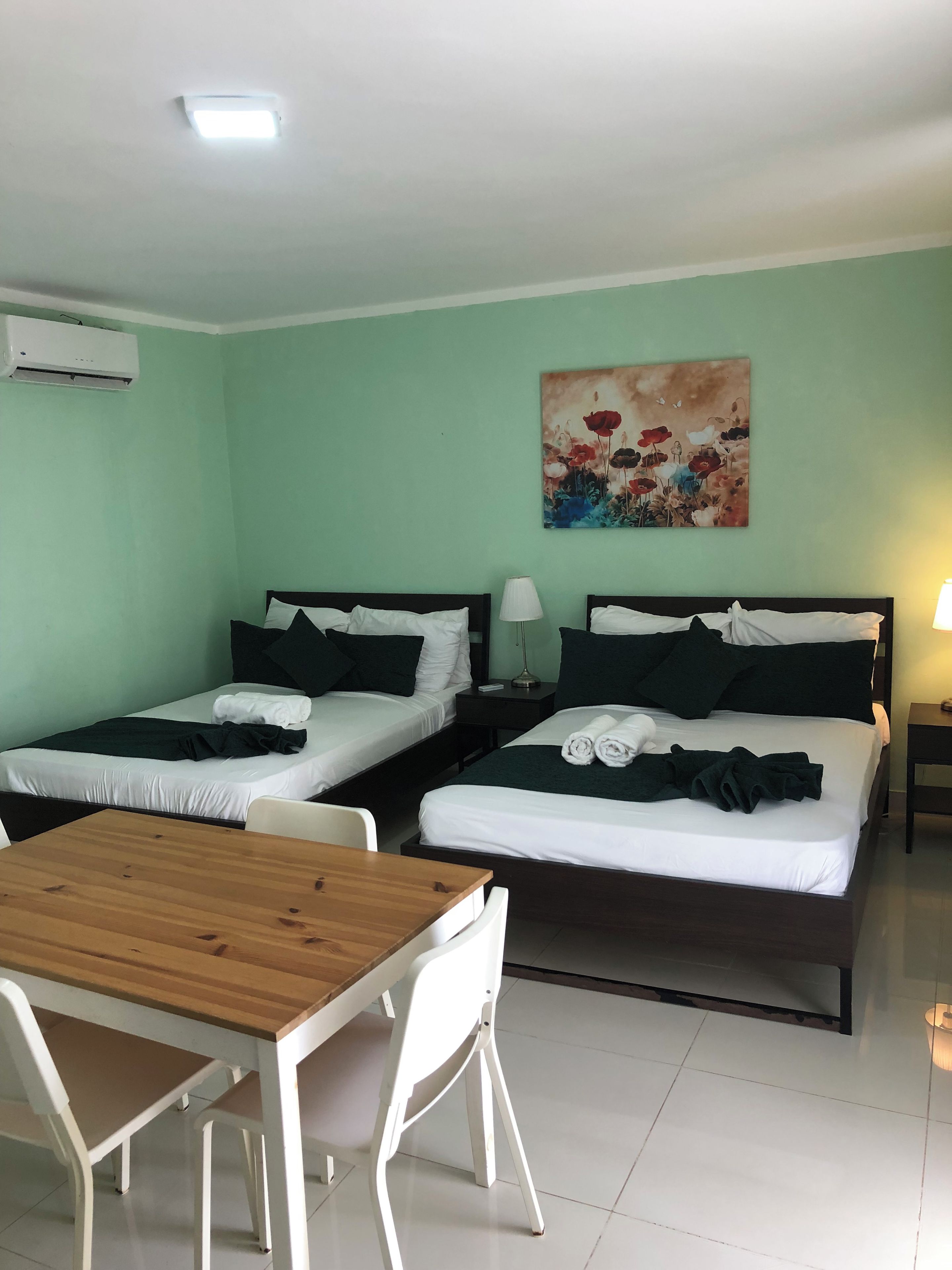 Une chambre d’hôtel avec deux lits simples, une table en bois, des chaises blanches, un tableau au mur et un appareil de climatisation.