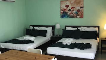 Une chambre d’hôtel avec deux lits simples, une table en bois, des chaises blanches, un tableau au mur et un appareil de climatisation.