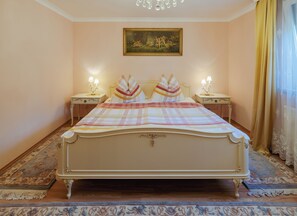Ein klassisches Himmelbett mit aufwendiger Verzierung, flankiert von Nachttischen mit Lampen, in einem Zimmer mit einem Blumenmuster-Teppich und einem gerahmten Gemälde an der Wand.