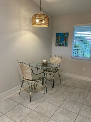 A dining area with a glass table and wicker chairs, a hanging light fixture, a painting on the wall, and a window with blinds. - Spacious 2-bedroom Condo with Golf course views in beautiful Naples (Naples)