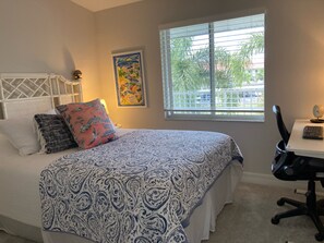 Une chambre à coucher comprenant un lit, un bureau avec une chaise, une fenêtre avec des stores et une œuvre d’art accrochée au mur.