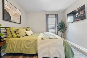 A bedroom with a bed, a bedside table, a window with curtains, a wall-mounted TV, and a plant. - Charming and inviting 3 bedroom - 1 bathroom home in Streamwood (Streamwood)