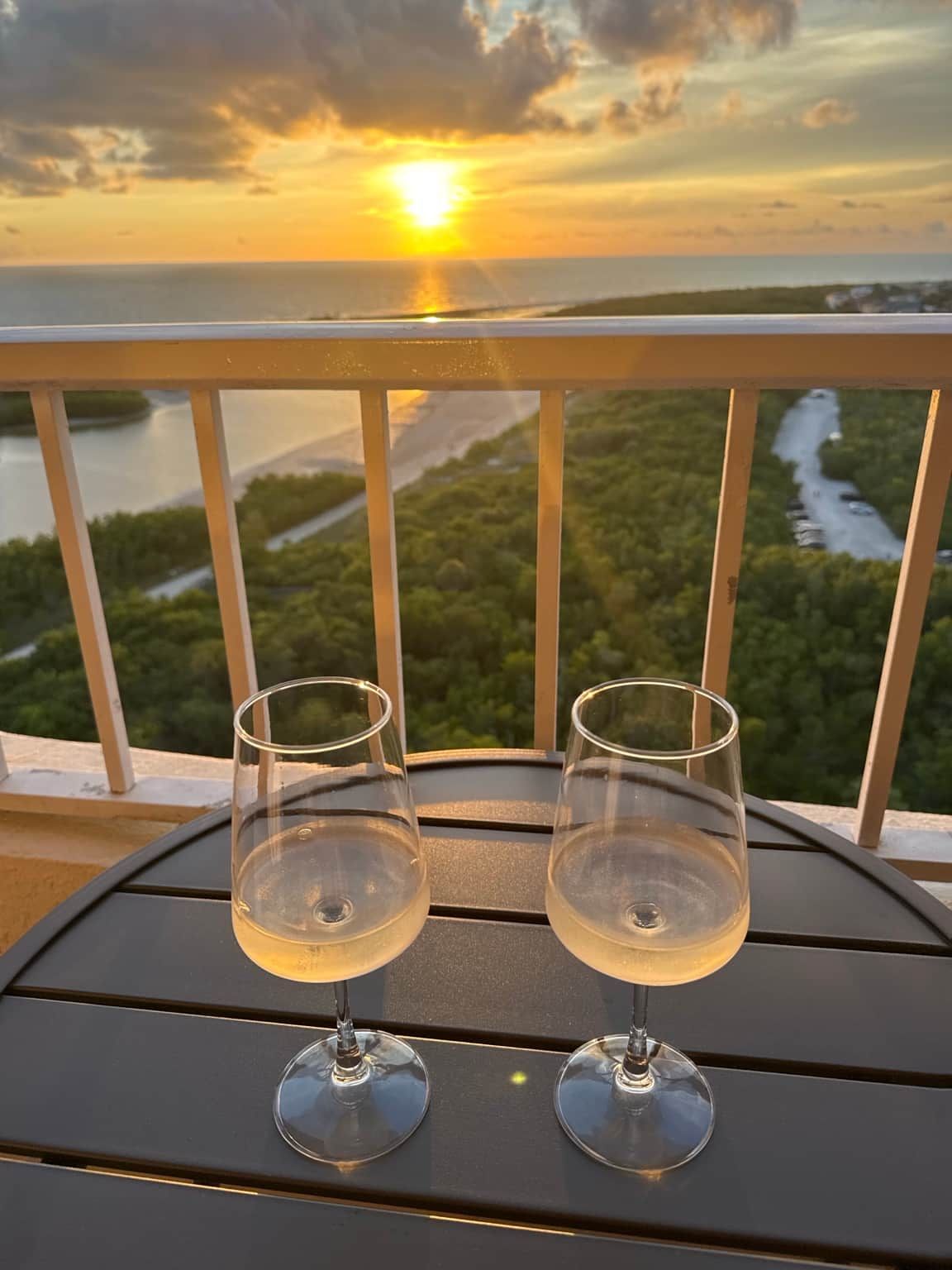 Two empty wine glasses on a table with a view of a sunset over a body of water and greenery.