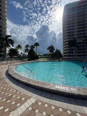 A swimming pool with a tiled deck, surrounded by palm trees and a multi-story building. - Sunsets at 19 Beachfront Penthouse Condo (Marco Island)