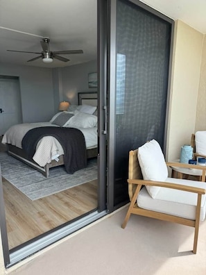 Une chambre avec un lit, un ventilateur de plafond, une porte-fenêtre coulissante et un coin salon comprenant une chaise et une petite table.
