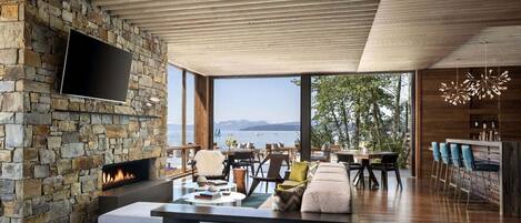 A modern living room with a stone fireplace, a flat-screen TV, and a view of the sea.