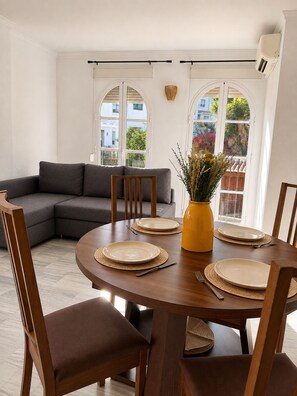 A dining area with a round wooden table set for four, surrounded by wooden chairs with brown cushions. A yellow vase with dried plants is on the table. - Montemar | 8-minute walk to the beach (Torremolinos)