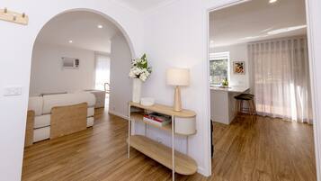 Sala de estar moderna com sofá, mesa de centro e abajur. Há um corredor dando acesso a outro quarto.