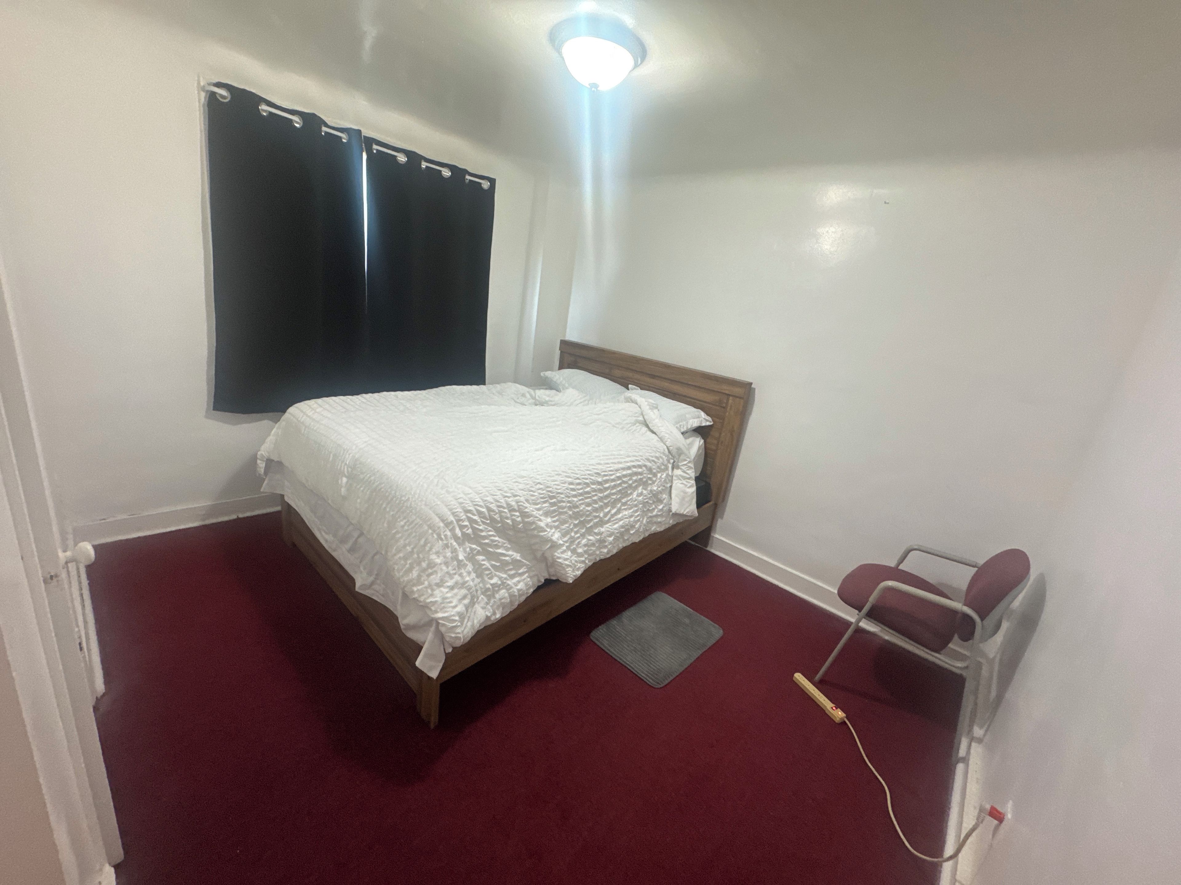 Un dormitorio con cama, ventana con cortinas, radiador y mesita de noche.