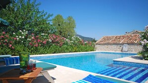 Piscina con sillas de descanso y una mesa, rodeada de plantas con flores y un muro de piedra.
