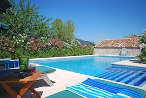 Piscina con sillas de descanso y una mesa, rodeada de plantas con flores y un muro de piedra.