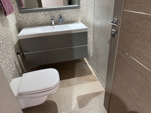 Une salle de bain avec des toilettes blanches, un lavabo surmonté d’un miroir et une porte située sur la droite.