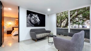 Uma sala de estar moderna com um sofá cinzento, uma poltrona cinza e um cobertor com padrão preto e branco. Um grande mural de tigre decora a parede, e o quarto possui uma grande janela com vista para árvores e uma varanda.