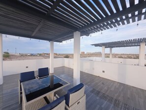 Pátio externo com mesa de vidro e cadeiras de vime, com vista para a paisagem desértica e edifícios ao longe.