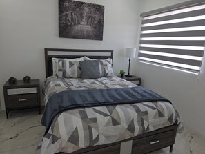 Quarto com cama, mesas de cabeceira, abajur, janela com persianas e quadro na parede.