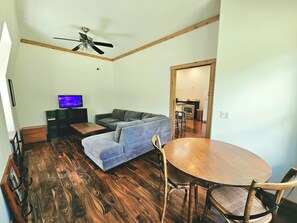 A living room with a grey sofa, a wooden coffee table, a television, and a dining area with a wooden table and chairs.