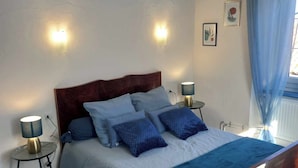 Un dormitorio con cama de madera, dos mesitas de noche, una mesita auxiliar, colcha azul y cortina azul.