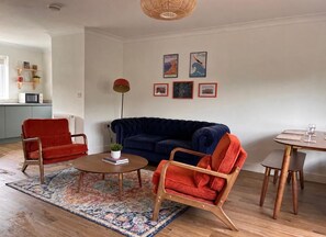 A living room with a dark blue sofa, a red armchair, a coffee table, and a dining area with a table and chairs. - The Bungalow Heacham (Heacham)