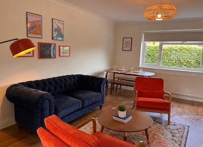 A living room with a dark blue sofa, a red chair, a round coffee table with a book and a plant, a dining table with chairs, and a window with a view of greenery. - The Bungalow Heacham (Heacham)