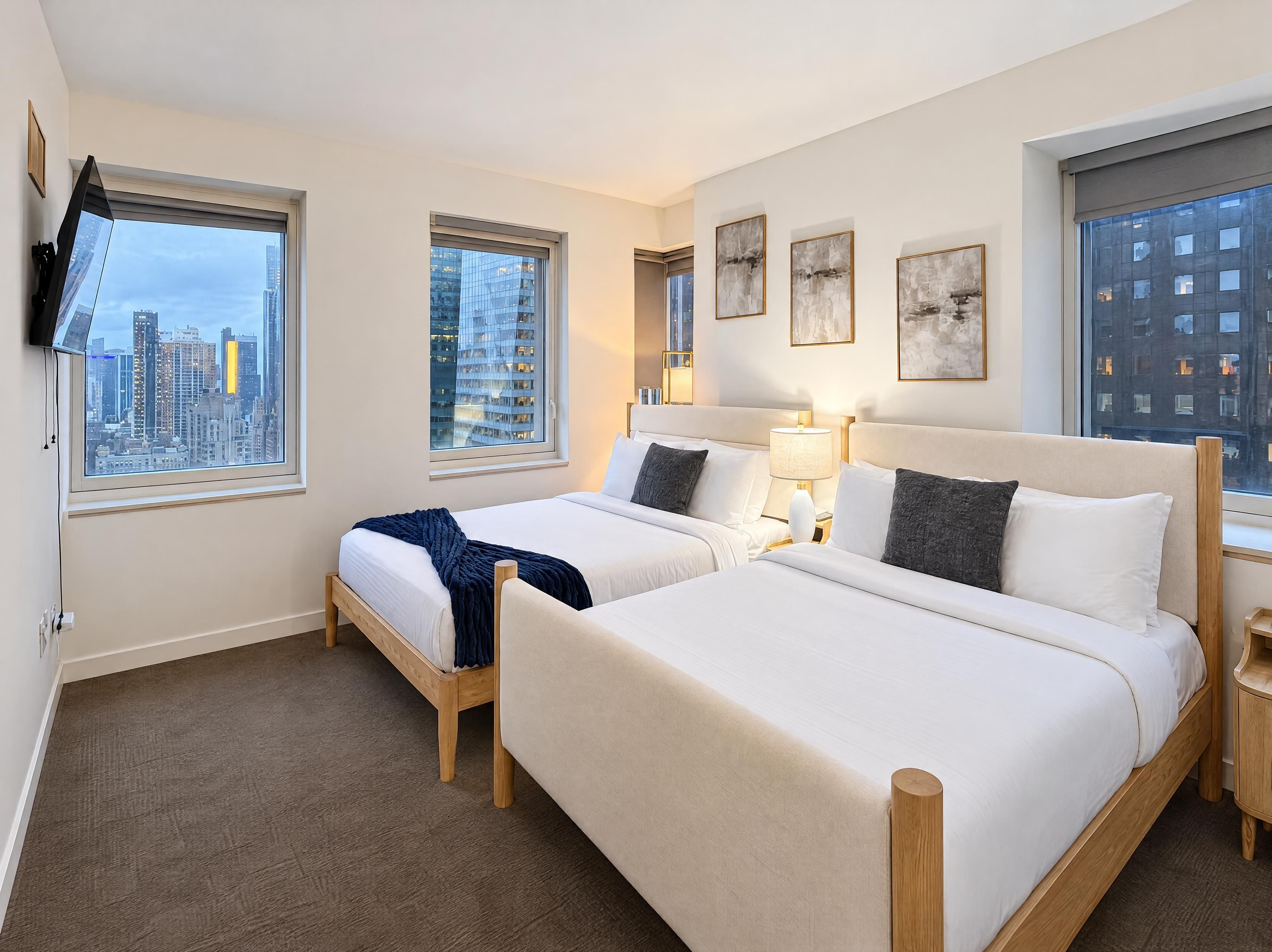 Ein Hotelzimmer mit zwei Betten, einem an der Wand montierten Fernseher und großen Fenstern mit Blick auf die Stadt.