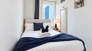 Een hotelkamer met een bed, een nachtkastje, een lamp, een raam met uitzicht op gebouwen en een aan de muur hangend kunstwerk.